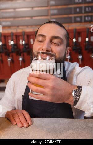 Vertikale Nahaufnahme eines Barkeepers, der fröhlich lächelt und frisch gebrautes Bier im Glas riecht Stockfoto