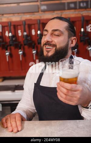 Vertikale Aufnahme von glücklichen bärtigen Barkeeper genießen Geschmack der Bier aus seiner Brauerei Stockfoto