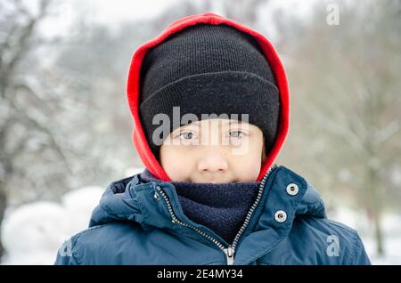 Porträt eines hübschen Jungen mit braunen Augen, der einen Mantel und eine Mütze trägt und draußen in einem verschneiten Park für eine Kamera posiert. Stockfoto