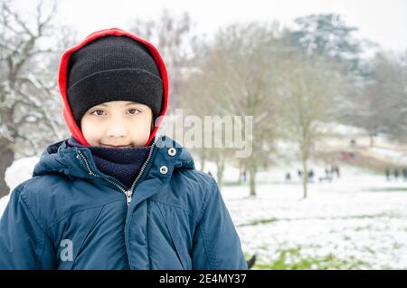 Porträt eines hübschen Jungen mit braunen Augen, der einen Mantel und eine Mütze trägt und draußen in einem verschneiten Park für eine Kamera posiert. Stockfoto