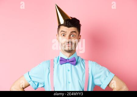 Feiertags- und Feiertagskonzept. Nahaufnahme eines verwirrten männlichen Entertainers in Partyhut und Fliege, verwirrt und schockiert, über Rosa stehend Stockfoto