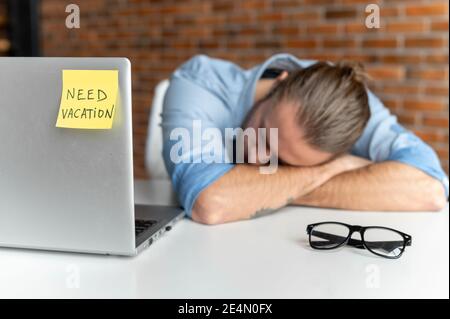 Ermüdete männliche Büroangestellte in smarter Freizeitkleidung macht ein Nickerchen am Arbeitsplatz, fühlt sich müde und Burnout, ein Hipster Kerl schläft auf dem Schreibtisch, ein Stockfoto