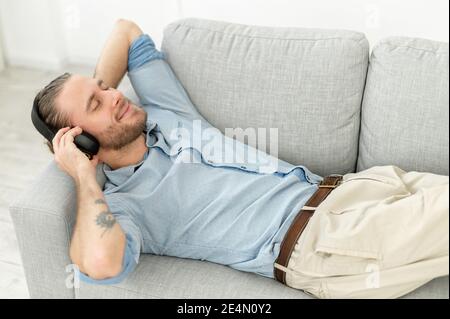 Fröhlicher gutaussehender Kerl mit kabellosen Kopfhörern zum Musikhören, ein Mann legt sich auf die Couch mit geschlossenen Augen verloren in Träumen, genießen Lieblings-Tracks Stockfoto