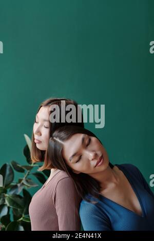 Junge Zwillingsschwestern asiatischer Ethnie mit dunklem, glattem Haar Entspannen vor der Kamera gegen Hausanlage mit Grün Blätter an Jadewand Stockfoto