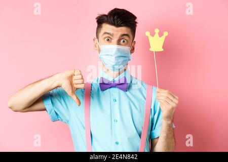 Covid, Pandemie- und Quarantänekonzept. Enttäuschter Typ in medizinischer Maske, der Partystrone und Daumen nach unten zeigt und sich beschwert, während er auf Rosa steht Stockfoto