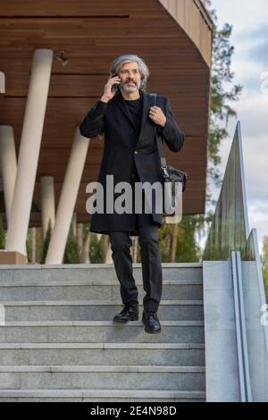 Selbstbewusster bärtiger grauhaariger Geschäftsmann mit Smartphone von Ear Consulting Client Oder einen Termin beim Umzug nach unten Stockfoto