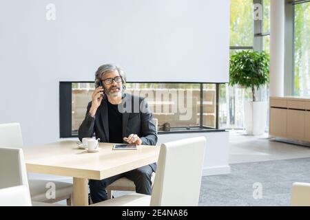 Moderner reifer Geschäftsmann in formeller Kleidung sitzt auf der Couch im luxuriösen Restaurant, spricht auf dem Smartphone und mit Touchpad Stockfoto