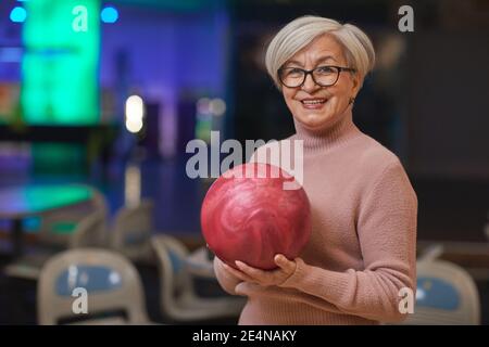 Waist-up-Porträt von lächelnden älteren Frau hält Bowling-Ball und Blick auf die Kamera, während aktive Unterhaltung in Bowling-Bahn genießen, kopieren Raum Stockfoto