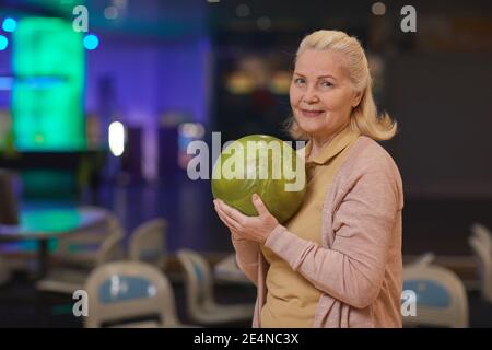 Waist-up-Porträt der eleganten älteren Frau hält Bowling-Ball und lächelt an der Kamera, während aktive Unterhaltung in Bowling-Bahn, Kopierer Platz genießen Stockfoto