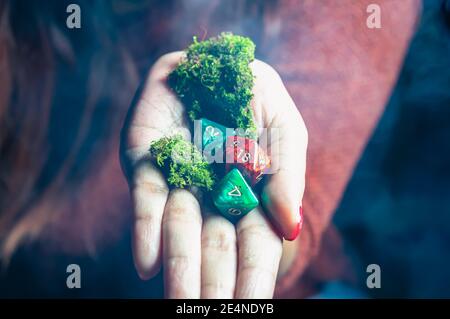 Ein roter 20-seitiger Würfel, zwei achtseitige Würfel und Moos in der Hand einer Frau in einer dunklen und rauchigen Umgebung Stockfoto