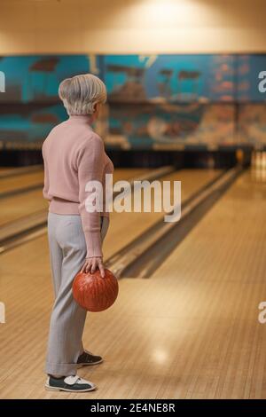 Vertikale Rückansicht Porträt der weißhaarigen älteren Frau spielen Bowling und wegschauen, während Sie an der Fahrspur halten Ball Stockfoto