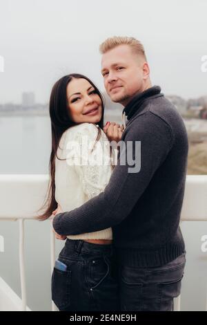 Liebespaar in Pullover kuschelt lächelnd auf der Brücke, im Hintergrund des Flusses und hohen Häusern. Hochwertige Fotos Stockfoto