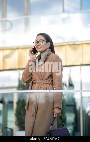 Lächelnde asiatische Geschäftsfrau in beigem Mantel im Gespräch auf Handy Während Sie im Freien vor dem modernen Business-Center stehen Stockfoto