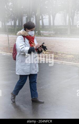 Selektiver Fokus auf einen nebligen Tag mit einer erwachsenen Frau Trägt eine Maske und trägt eine Tasche auf dem Rücken Gehen und Verwenden eines Mobiltelefons zum Senden und Empfangen Stockfoto