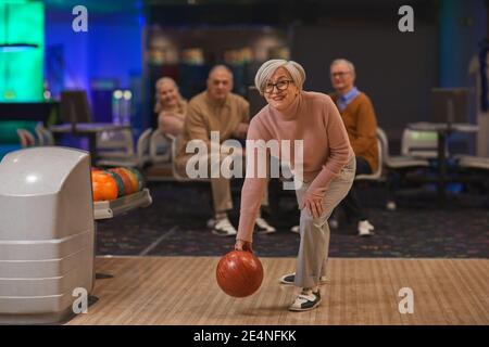 In voller Länge Porträt der freudigen älteren Frau Bowling mit Gruppe von Freunden im Hintergrund spielen, Raum kopieren Stockfoto