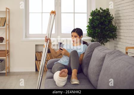 Frau mit gebrochenem Bein mit Handy, lesen Nachrichten online und Leiden allein, nicht in der Lage zu gehen Stockfoto