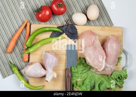 Rohe Hähnchenschenkel und Filet auf Holzbrett mit Messer, Eiern und frischem Gemüse Stockfoto