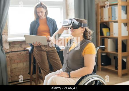 Junge deaktivieren weiblich in vr Headset im Rollstuhl sitzen und Präsentation ansehen, während ihre Kollegin ihren Laptop am Arbeitsplatz verwendet Stockfoto