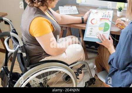 Zwei junge Makler in Casualwear diskutieren Finanzcharts und Diagramme, während einer von ihnen auf Daten zeigt und erklärt IT Stockfoto