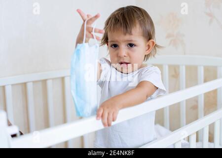 Coronavirus covid-19 Quarantänekonzept, Vorsichtsmaßnahmen. Kleines niedliches Kleinkind steht in der Krippe zu Hause und hält eine medizinische Maske. Stockfoto