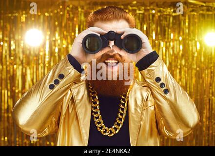 Fröhlicher lustiger Mann in einem funky Outfit mit einem riesigen Goldene Kette, die durch ein Fernglas schaut Stockfoto