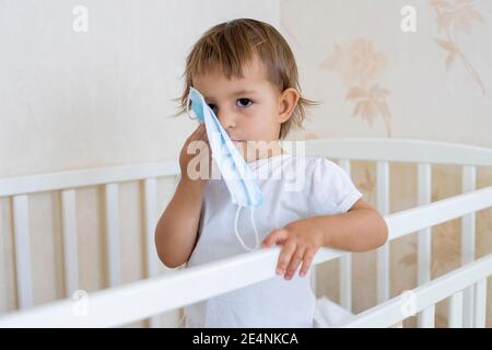 Concept Coronavirus Lockdown. Kleine niedliche Kleinkind steht in der Krippe zu Hause und umarmt eine medizinische Maske Stockfoto