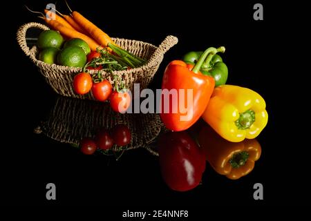 Stillleben mit roten, grünen und roten Paprika in einem schönen Weidenkorb auf schwarzem Hintergrund mit Reflexion Stockfoto