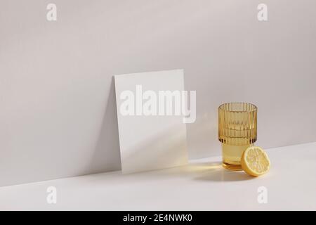 Sommer Schreibwaren Stillleben Szene. Schneiden Sie Zitronenfrucht und goldenes gewelltes Glas Wasser auf beigem Tischhintergrund in Sonnenlicht. Leere Papierkarte Stockfoto