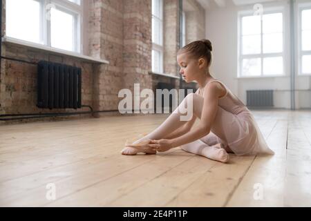 Kleine Ballerina Mädchen sitzt auf dem Boden des Tanzstudios Und die spitzenschuhe vor dem Unterricht anziehen Stockfoto