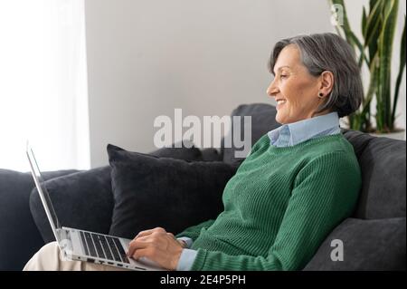 Eine entspannte ältere Dame in einem grünen Pullover chillt im Wohnzimmer, bequem auf der Couch oder Sofa sitzen, eine Comedy-Show auf der zu sehen Stockfoto