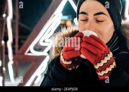 Junge kaukasische Mann trinken heiße Schokolade, Kaffee oder Tee in Winterkleidung und rote Handschuhe vor einem Hintergrund von fluoreszierenden Hintergrundbeleuchtung Stockfoto