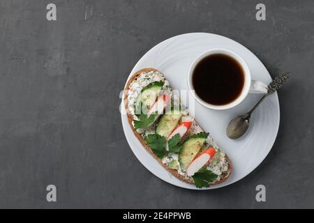 Hausgemachtes Sandwich mit Krabbenstäbchen, Gurke, Frischkäse und einer Tasse Kaffee auf dunklem Hintergrund. Blick von oben Stockfoto