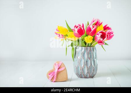 Recycelte herzförmige Geschenkbox mit Satinband Schleife und Frühlingsstrauß von rosa und gelben Tulpenblüten in einem Vase auf dem weißen Holztisch und weiß Stockfoto