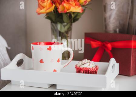 Tablett mit frischem Kaffee und Cupcake, Geschenkbox und einem Strauß Rosenblumen in Vase auf dem Nachttisch gegen eine graue Wand. Valentinstag Stockfoto