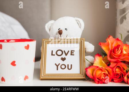 Morgen Tasse frischen Kaffee, Rosen Bouquet, weißen Teddybär mit Liebe Sie Nachricht im Rahmen auf dem Nachttisch. Valentinstag, Geburtstag, Frauen Stockfoto