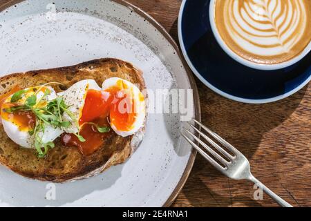 Eine Tasse Kaffee mit Latte Art drauf, geröstetes Sauerteig-Brot mit pochiertem Ei auf dem Tisch im Café. Stockfoto