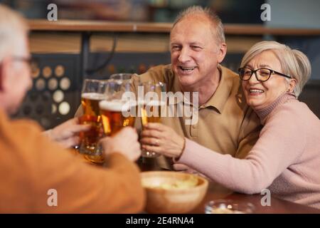 Porträt eines lächelnden älteren Paares, das in der Bar und Bier trinkt Klirrende Gläser, während Sie die Nacht mit Freunden genießen Stockfoto