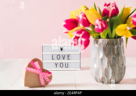 Lightbox mit Liebe Sie Nachricht und verschwommen Herzform Geschenk Box und Frühling Bouquet von rosa und gelben Tulpenblüten In einer Vase auf dem weißen Holz Stockfoto