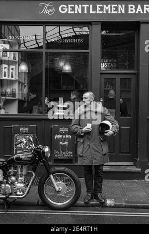 Ein Mann macht eine Kaffeepause in einem Café London mit klassischem Motorrad vor einem Kaffee geparkt Kaufen Stockfoto