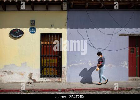 Frau mit Maske auf dem Bürgersteig vor bunten Häusern und Geschäften in Antigua, Guatemala, Mittelamerika. Stockfoto