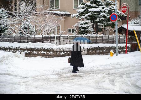 Ein Mann, der einen langen schwarzen Mantel trägt und einen blauen Regenschirm hält, der seinen Weg durch eine schneebedeckte Straße in einer japanischen Stadt bahnt. Stockfoto