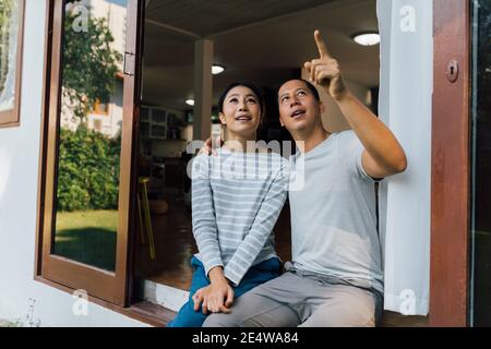 Portrait von jungen erwachsenen asiatischen Paar suchen nach oben, während ein Mann zeigt mit Finger in Hause inneren Hintergrund. 30s reifen erfolgreichen Ehemann und Ehefrau. Immobilien- und Immobilienleistungskonzept Stockfoto