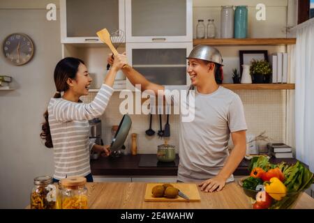 Junge verheiratete Paar tun einen vorgeblichen casual Kampf mit Utensilien und Pfannen in der Küche zu Hause. Lustige Mann und Frau halten ein Küchengeschirr mit einer lustigen Zeit Stockfoto