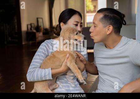 Junge Erwachsene asiatische Paar mit einem Welpen mit Zuhause Interieur im Hintergrund. 30s reifer Mann küssen einen Hund Haustier in den Armen der Frau. Mann und Frau leben zusammen im Haus. Modernes Familienglück Stockfoto