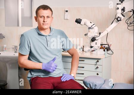 Vorderansicht des jungen Mannes Zahnarzt in sterilen Handschuhen Blick auf die Kamera und zeigt Genehmigung Geste. Männlicher Stomatologe in blauem Hemd sitzt in der Nähe von zahnmedizinischen diagnostischen Mikroskop. Konzept der Zahnmedizin. Stockfoto