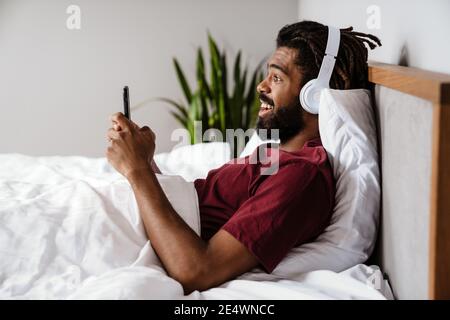 Aufgeregt afroamerikanischen Mann in Kopfhörer mit Handy während liegen Im Bett zu Hause Stockfoto