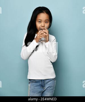 Positive asiatische koreanische Mädchen in Jeans und weißen Langarm Stehen und trinken reines Wasser aus Glas auf blauem Hintergrund Stockfoto