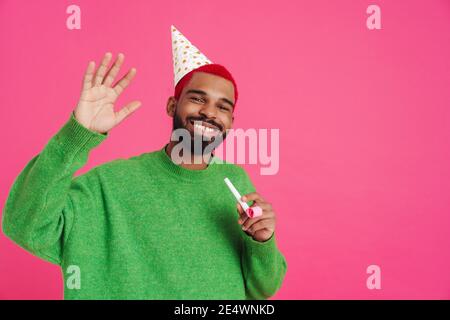 Fröhlicher afroamerikanischer Kerl winkt Hand und hält Party Pfeife Isoliert auf rosa Hintergrund Stockfoto