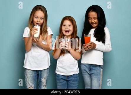 Glücklich lachende Mädchen Freunde Kinder in weißen T-Shirts stehen mit Wasser, Milch oder Kefir und frischen Karottensaft Stockfoto
