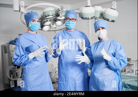 Vorderansicht von drei Ärzten im Operationssaal, Chirurgen bereit für die plastische Chirurgie in der Klinik, tragen sterile Handschuhe, blaue OP-Uniform, schützende Gesichtsmaske. Konzept der Medizin Mitarbeiter im Gesundheitswesen Stockfoto